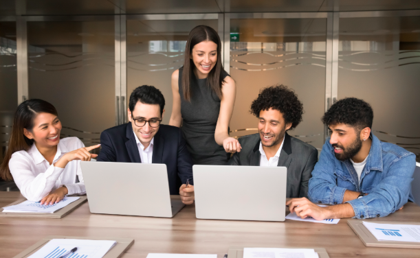 A diverse group of business professionals in a meeting looking at a couple of laptops while smiling