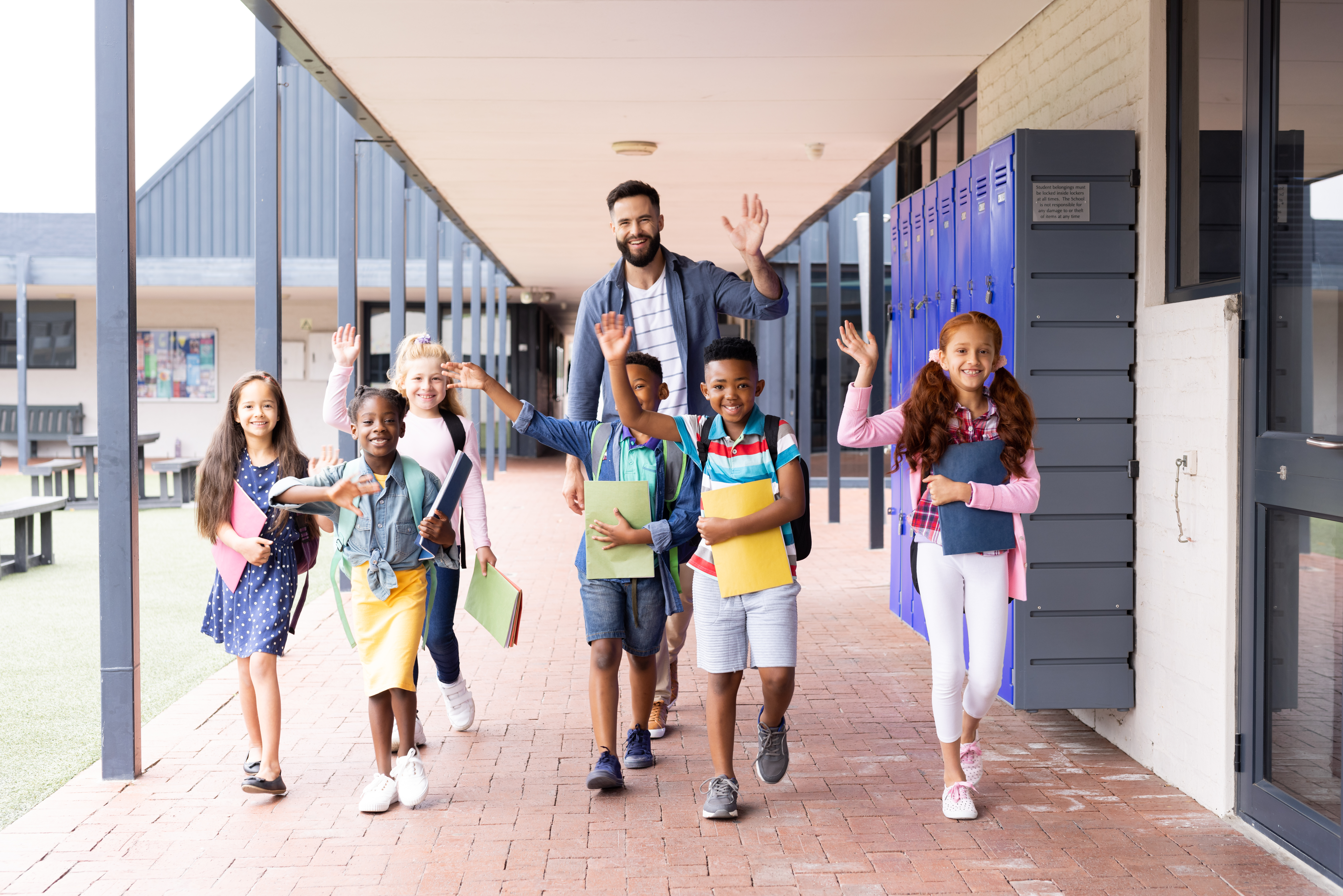 happy male elementary teacher and diverse schoolchildren smile and wave at the camera